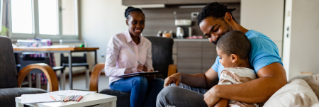 A practitioner sits across from a father and his child in a living room. The practitioner smiles and takes notes.