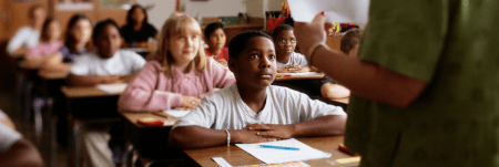 A classroom of students sit at their desk with a pencil and paper in front of them, looking up at their teacher.