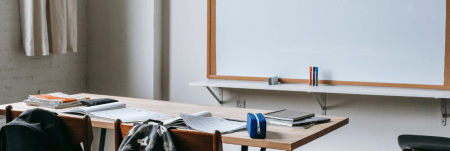 An empty classroom with a whiteboard, a desk and a couple of chairs placed in the front. The desk is occupied by student supplies.