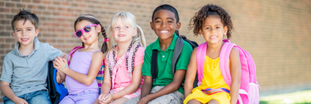 A group of children sit next to each other with their backpacks on and smile.