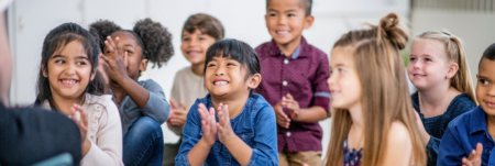 A group of children sit together facing their teacher. They smile and clap their hands.