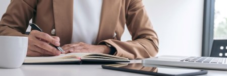 A person sitting at their desk, writing in their notebook, with their laptop and tablet in front of them.
