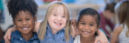 Three young girls smile and pose for a photo. The girl in the middle has her arms around the two girls beside her.