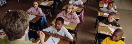 A classroom of students sit at their desk, attentively listening to their teacher.