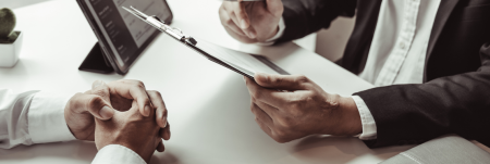 Two professionals sit at a table. One person holds up a clipboard, going over information.