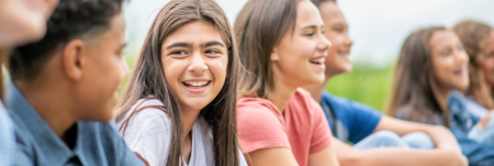 A group of children sitting on grass. Two of the kids look at each other smiling, while the others look forward and smile.