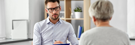 A practitioner is sitting down with their patient, attentively listening and writing down information on a clipboard.
