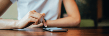 A person sitting at a table, resting their forearms, and viewing their phone.
