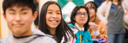 Students sitting at their desks in a row in a classroom. One student is smiling.