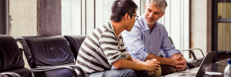 Two individuals casually sit at a conference table. They face each other in a one-on-one conversation.