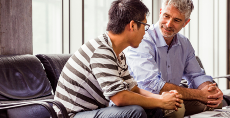 Two individuals casually sit at a conference table. They face each other in a one-on-one conversation.