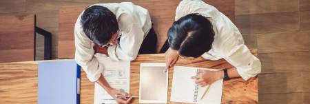 An overhead view of two individuals sitting around a table with a tablet and papers.