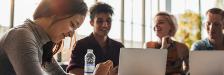 A group of young adults are working together at a table with their laptops open. They are smiling and laughing.
