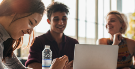 A group of young adults are working together at a table with their laptops open. They are smiling and laughing.