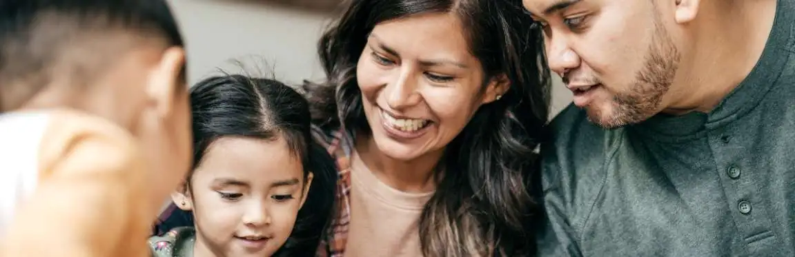 A family sits close together. The mother and father watch the children play as the mother smiles.