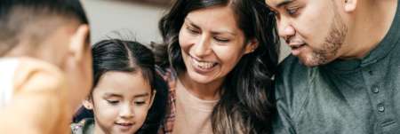 A family sits close together. The mother and father watch the children play as the mother smiles.