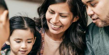 A family sits close together. The mother and father watch the children play as the mother smiles.