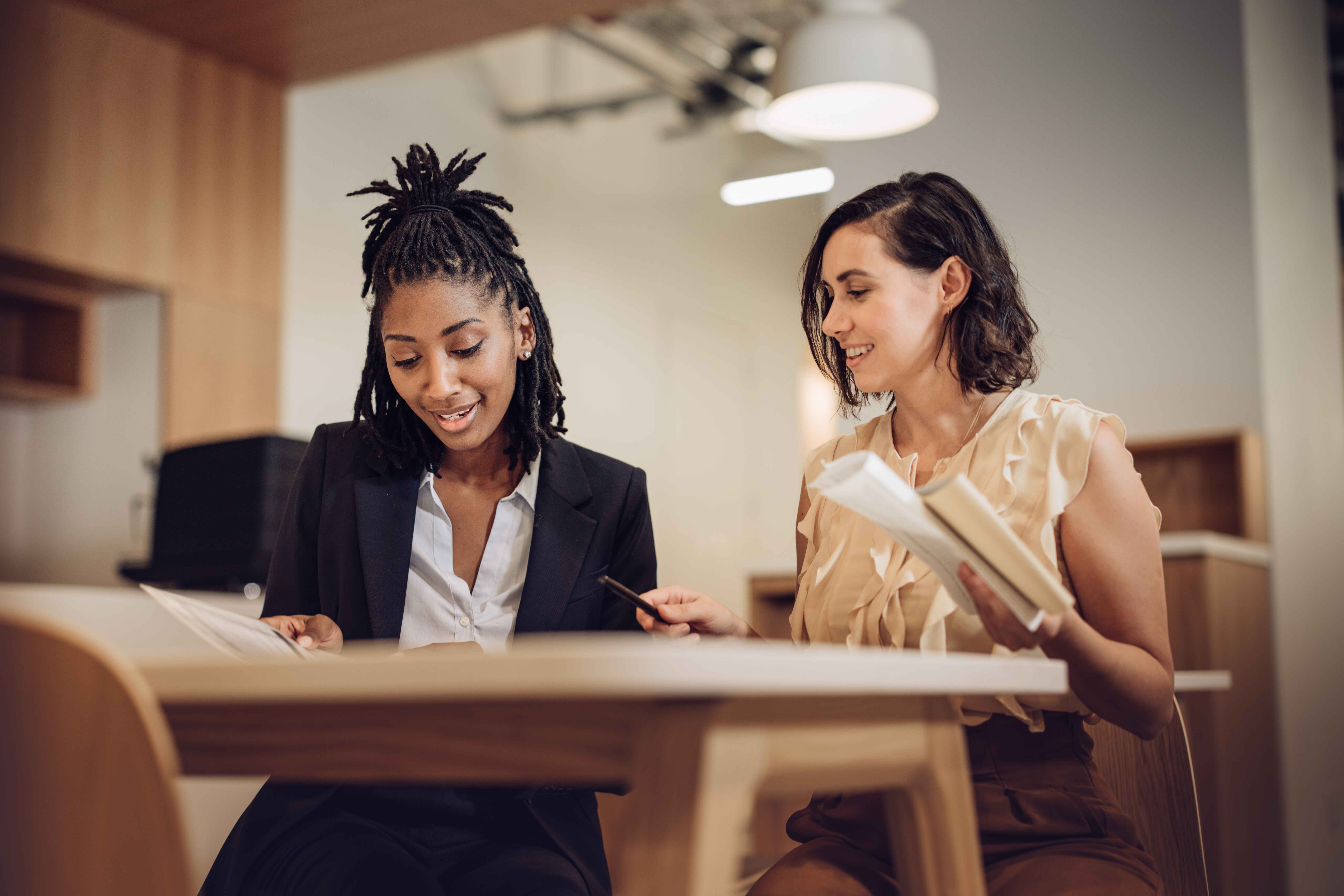 Two people sit together at a table in a modern office space. They both look down at the table and smile.