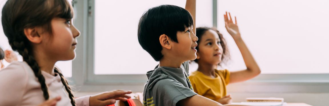 Three young students sitting at their desks in a classroom. Two students have their hand raised.