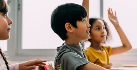 Three young students sitting at their desks in a classroom. Two students have their hand raised.