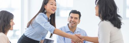 A sales professional is standing and shaking another person's hand across a table. Two other people are seated at the table.