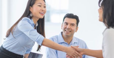 A sales professional is standing and shaking another person's hand across a table. Two other people are seated at the table.