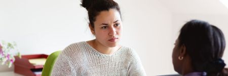 Two people sit across from each other, having a conversation. There is a green chair and flowers in the background.
