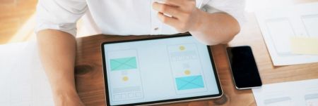 A person working on a tablet with mobile app wireframes at a desk.