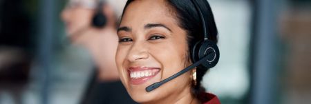 A person smiling, wearing a headset with a microphone, working in a customer service environment.