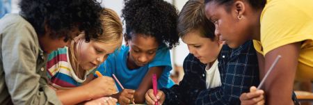 A group of young students standing over a desk in a classroom setting and writing on a poster.