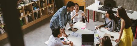 A group of six people are gathered around a white table in an office setting. The table is cluttered with laptops, notebooks, papers, sticky notes, and coffee cups. One person is standing and leaning over the table while the others are seated or standing around it. Shelves filled with books and other items line the wall in the background. Another desk with a computer setup is visible in the background as well.