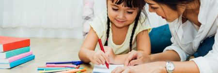 An adult and a child sit on the floor drawing together. The child uses a red pencil while colored pencils and books lie nearby.