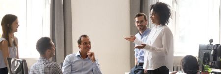 A group of five people are having a discussion in an office setting. One person is standing and gesturing with their hand, while the other four are seated or standing around a table.
