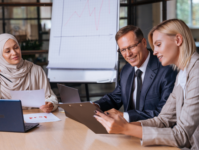 Three people sitting at a table with laptops and documents, discussing business matters with a flip chart showing graphs in the background.