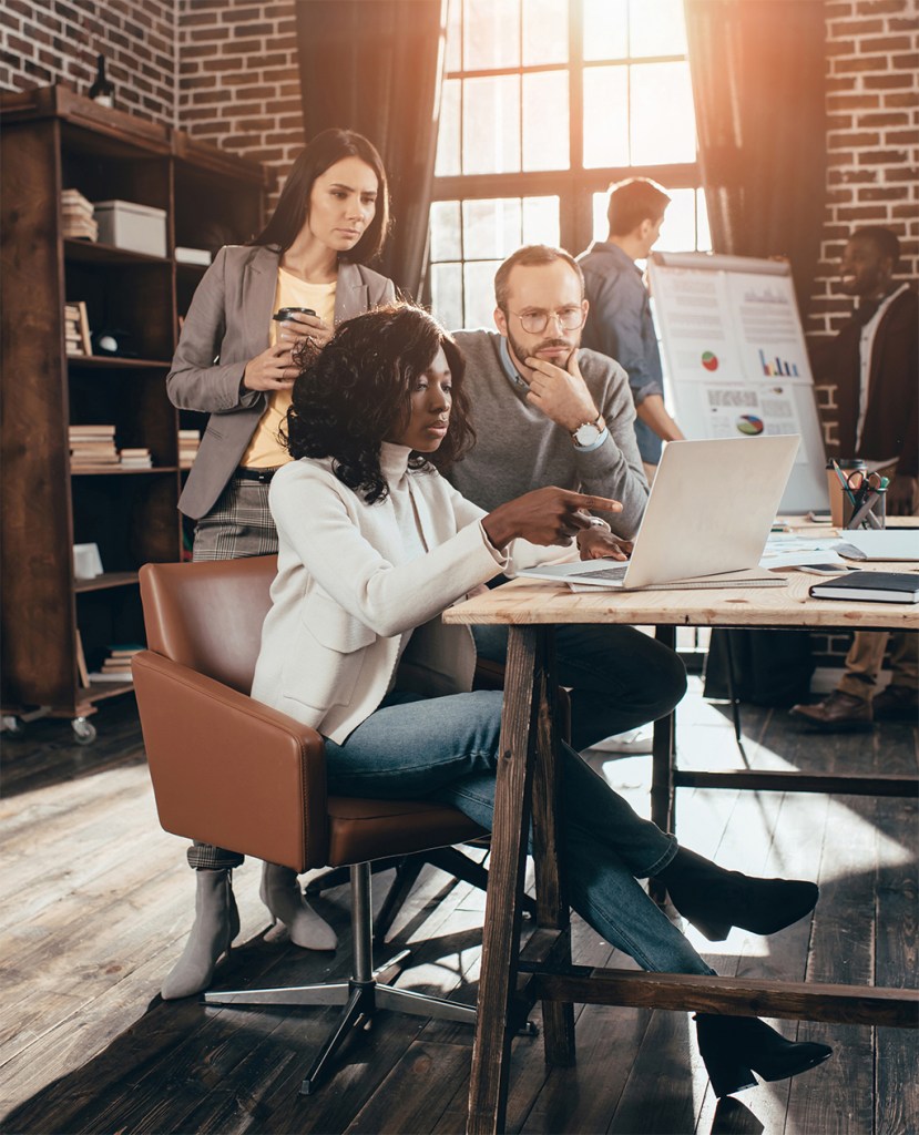 Five team members sitting around a conference table reviewing architectural plans and documents during a brainstorming session led by one individual pointing at a blueprint.
