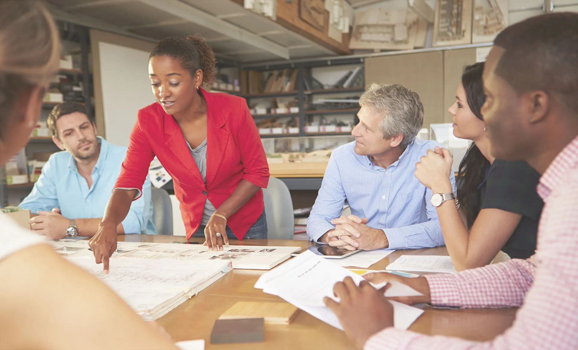 A group of five people are gathered around a table in an office setting. One person, wearing a red blazer, is standing and pointing at documents spread out on the table while speaking to the others. The other four individuals are seated and attentively listening, with papers and notebooks in front of them.