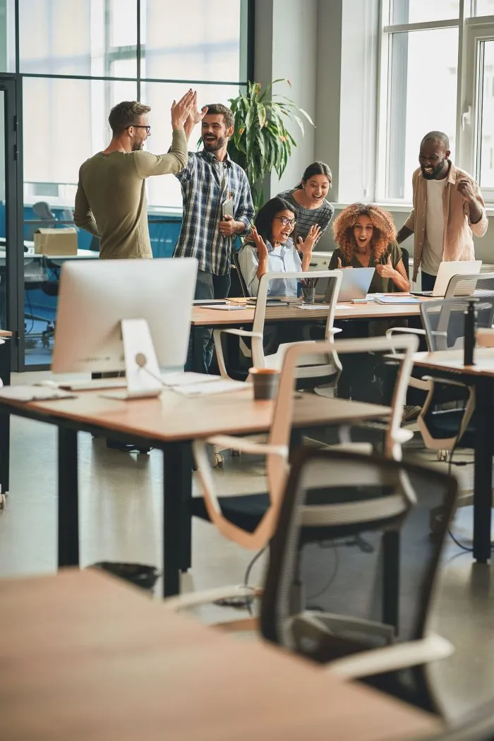 Six people in an open office space celebrating with high-fives and cheering around desks equipped with computers and office supplies.