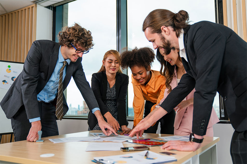 Five individuals standing around a table covered with papers and charts, actively discussing documents in a well-lit office with large windows.