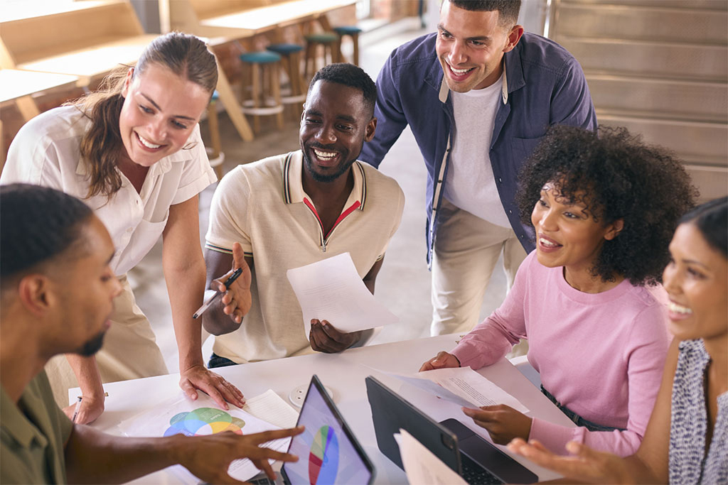 Six individuals gathered around a table, engaging in conversation while reviewing documents and laptops in a casual professional setting.