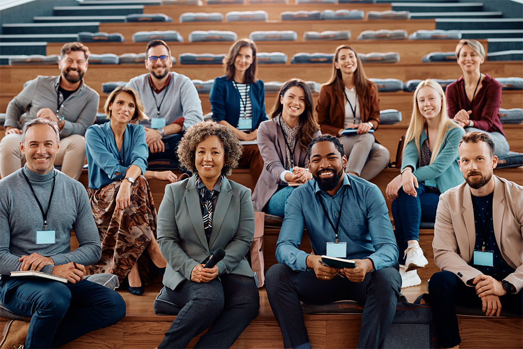 A group of twelve people seated on tiered benches in an auditorium or lecture hall, posing for a group photo with microphones or notepads.