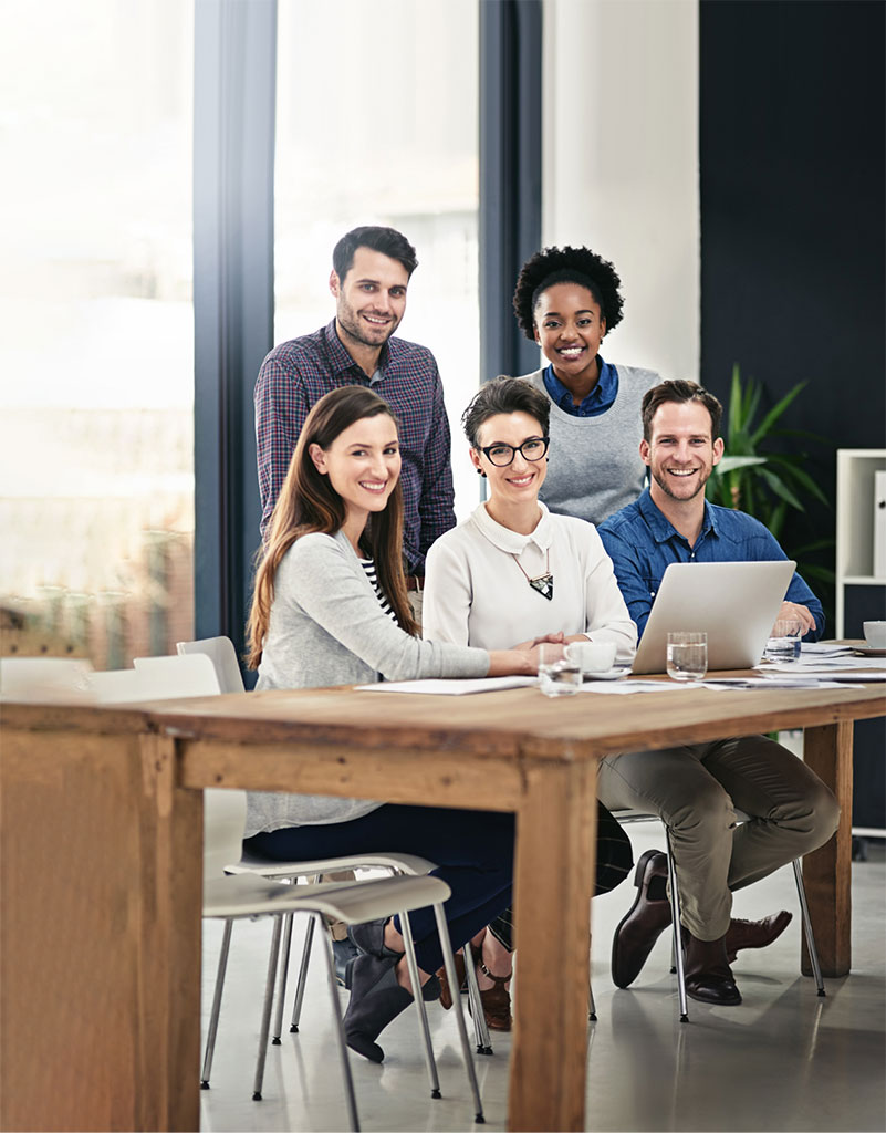 Five people sitting around a wooden table working on laptops and discussing together in an informal meeting room or workspace.