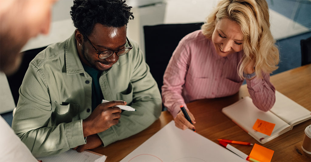 Two individuals working together at a desk covered with papers, notebooks, sticky notes, and coffee cups; one writing while the other observes.