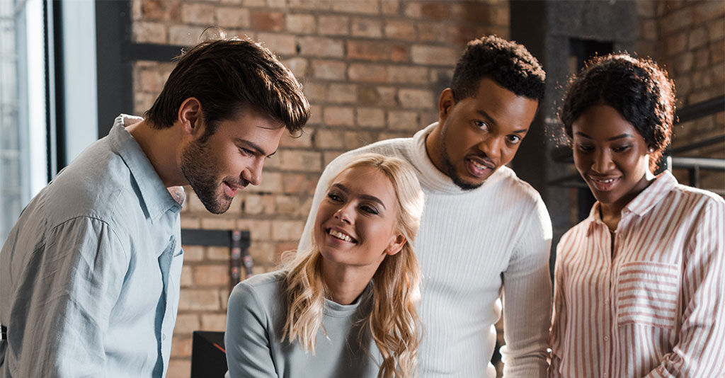 Four coworkers collaborating on a project in an industrial-style workspace with exposed brick walls and large windows, focused on a laptop screen.