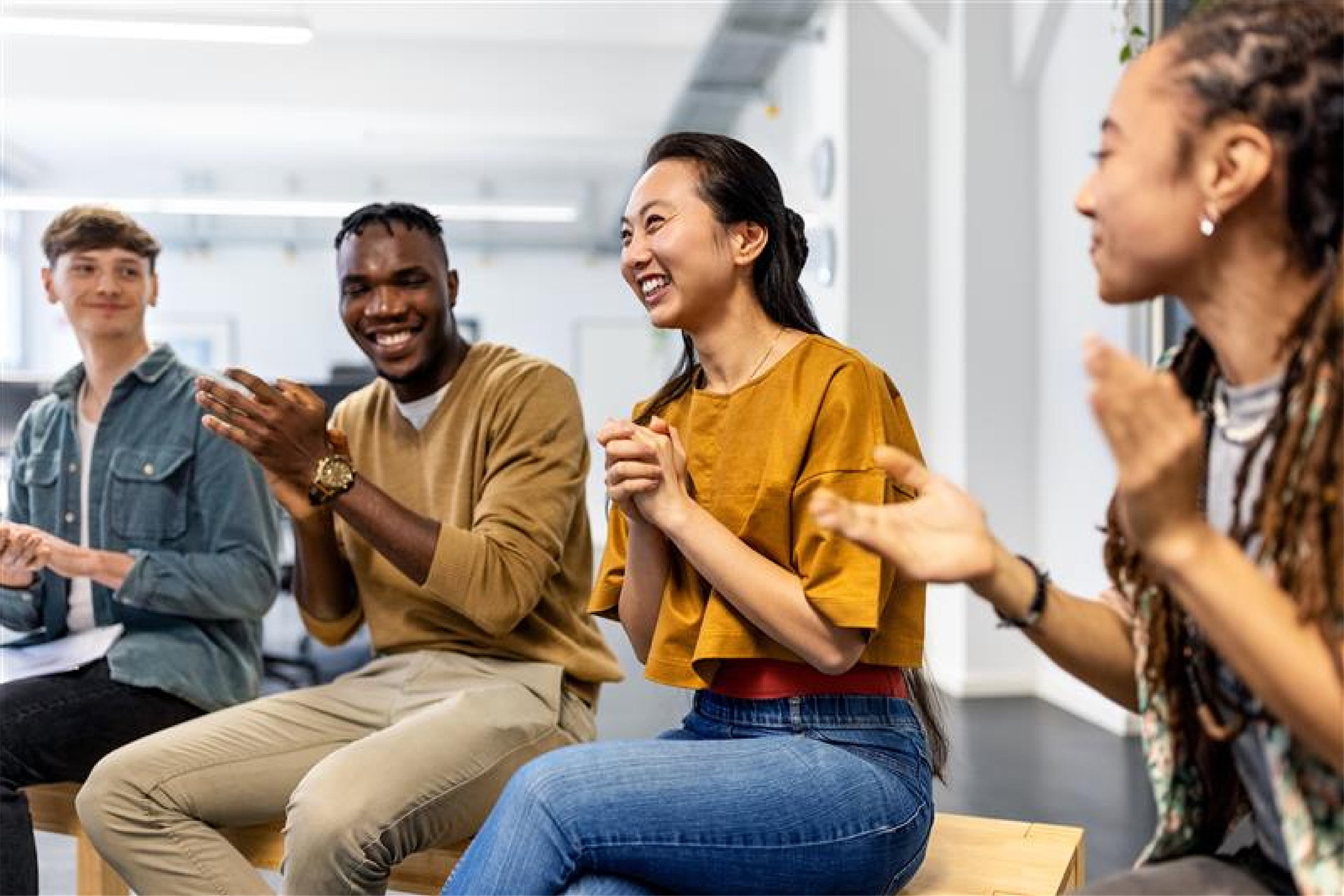 Four people sit in a bright room with large windows, clapping and smiling during an informal meeting or discussion.