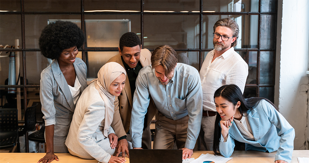 Six people gathered around a table in an office, looking at a laptop and discussing something together.